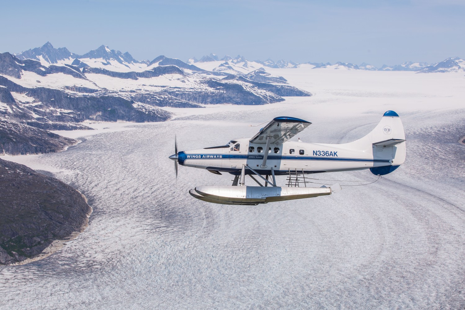 Seaplane flying over glacier with snow-capped mountains in the background.